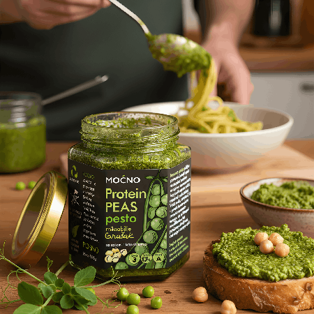 Serving view: a spoon scoops pesto spread from the jar, while in the background there is a plate of pasta and toasted bread with the spread, showing the culinary application of the product.